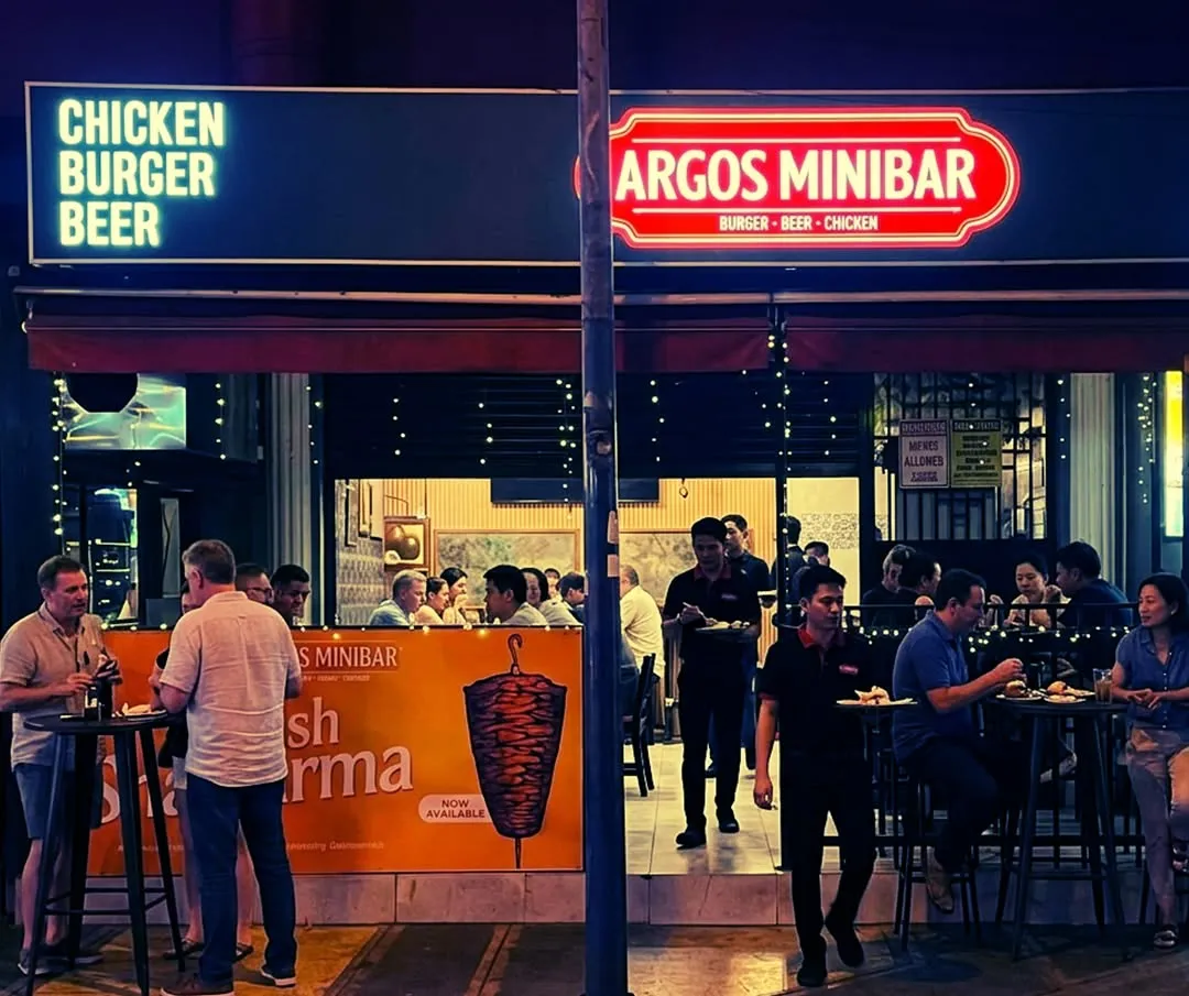 Argos Minibar's open-front dining area lit at night with guests seated inside.