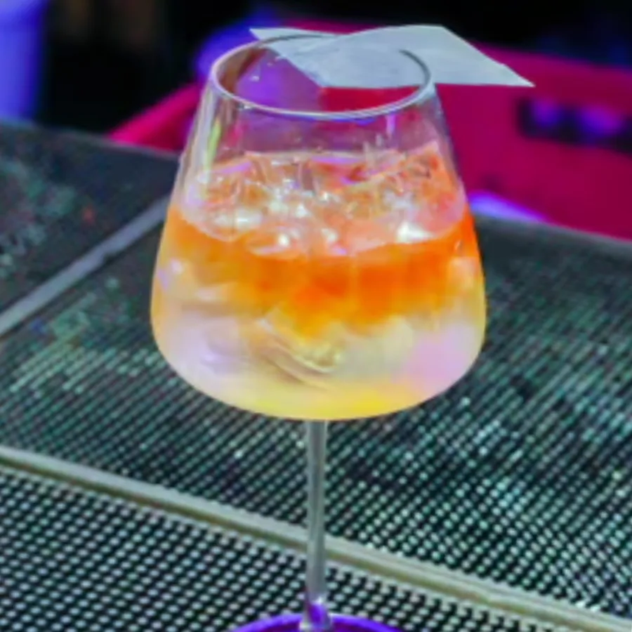 Orange layered cocktail in a stemmed glass under colored bar lights.