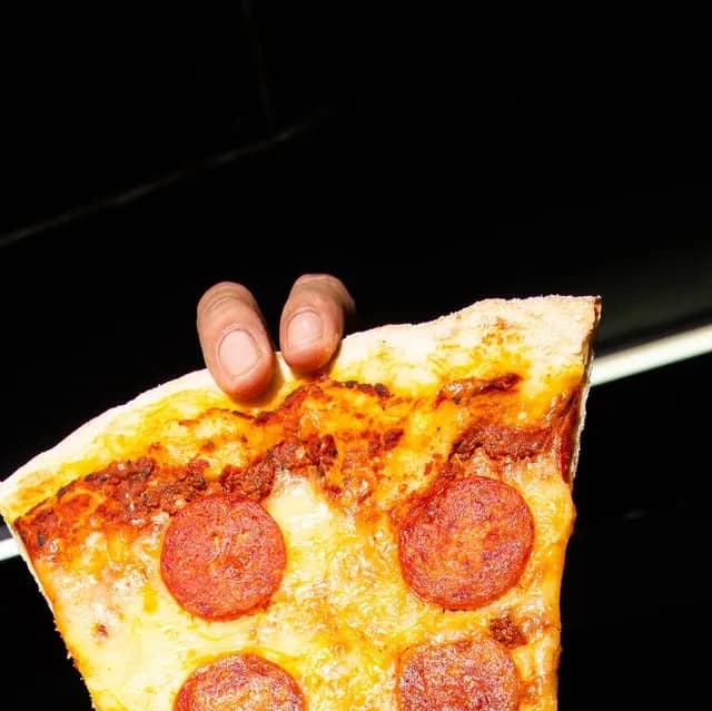 A hand holding a large pepperoni pizza slice against a dark background.