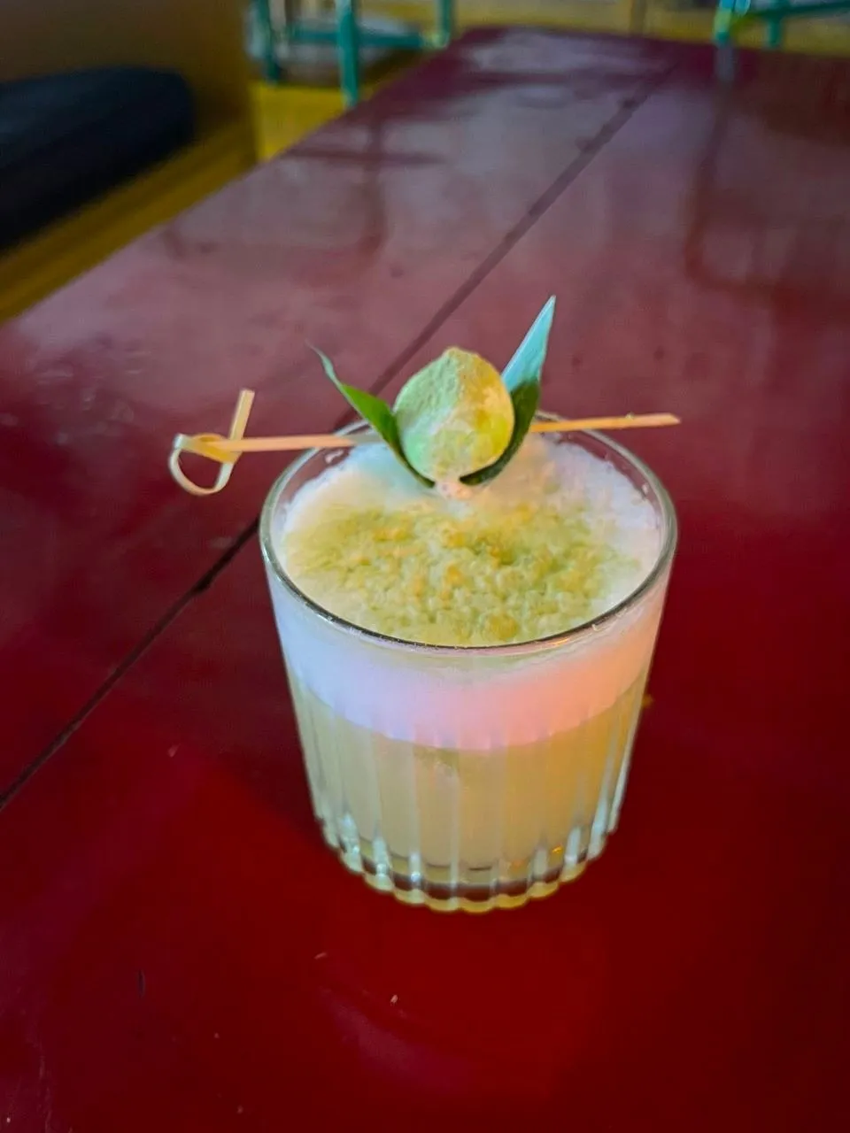 A pale foamy cocktail with a calamansi garnish on a red table.