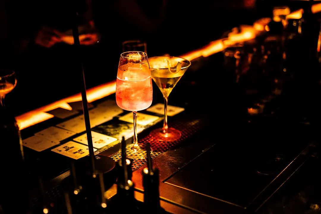 Two cocktails glowing on the bar in Cheshire's dark room.