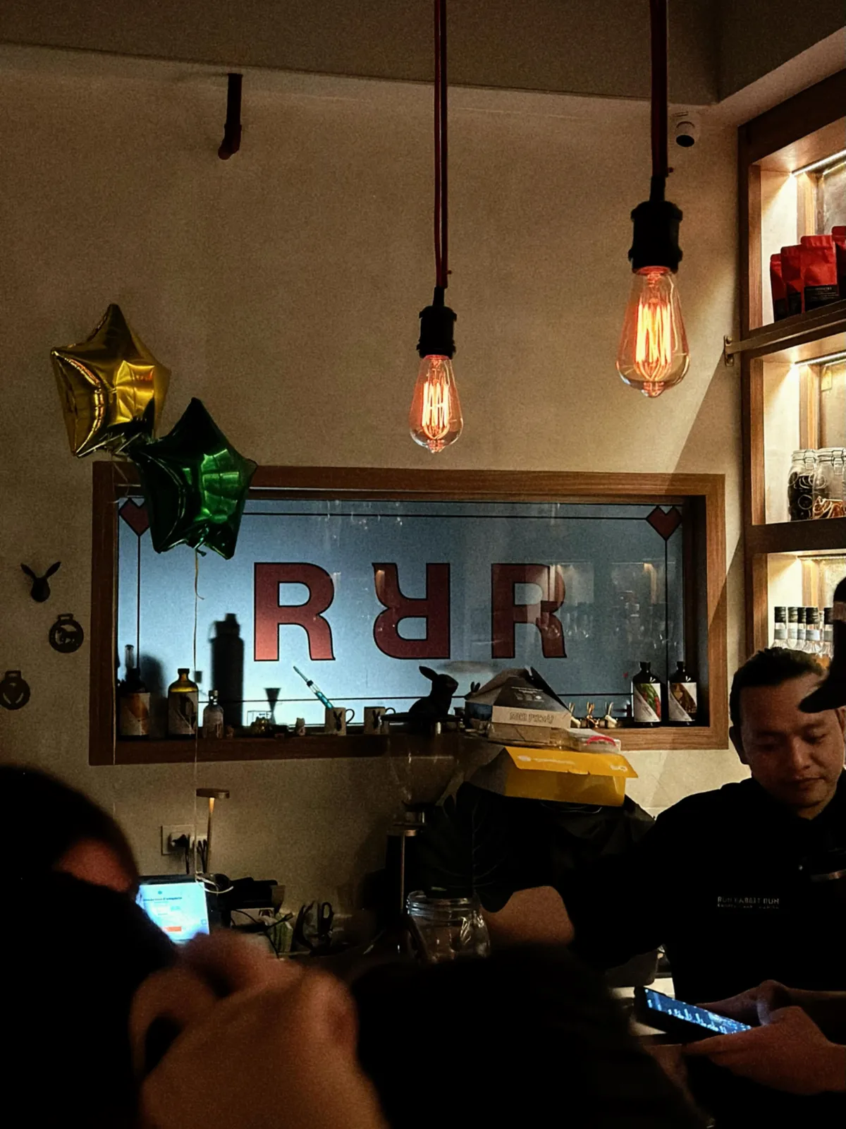 Warm bar room with hanging lights, a window into the service area and guests in the background
