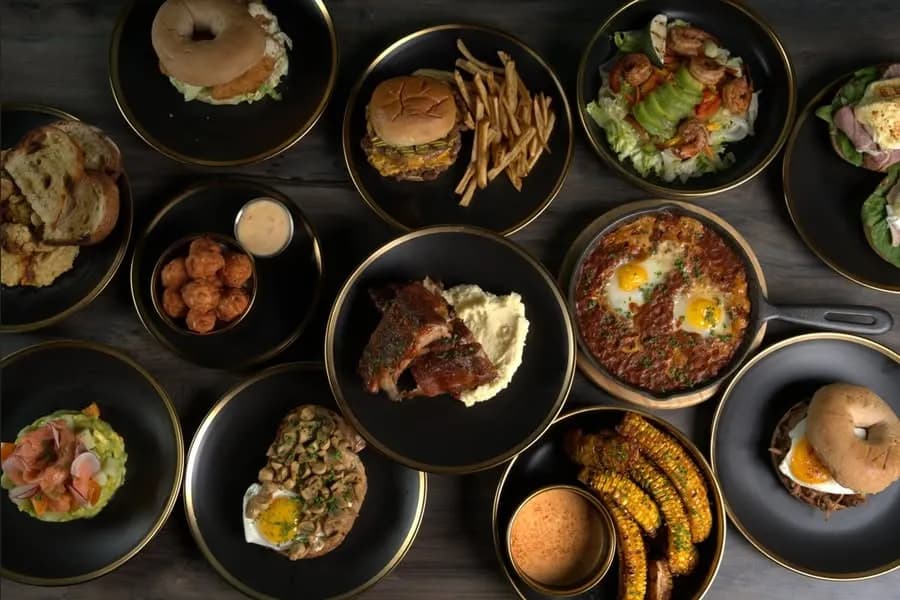 Overhead spread of burgers, wings, chicken, corn, salad and bagel dishes.