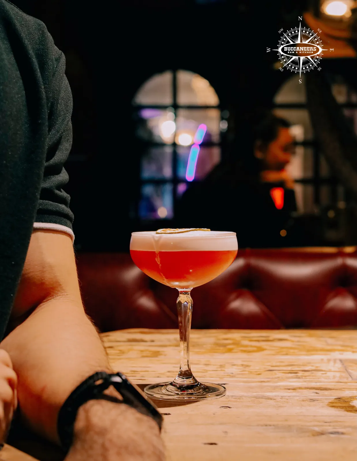 Red cocktail in a coupe on a wooden table inside the dim bar.