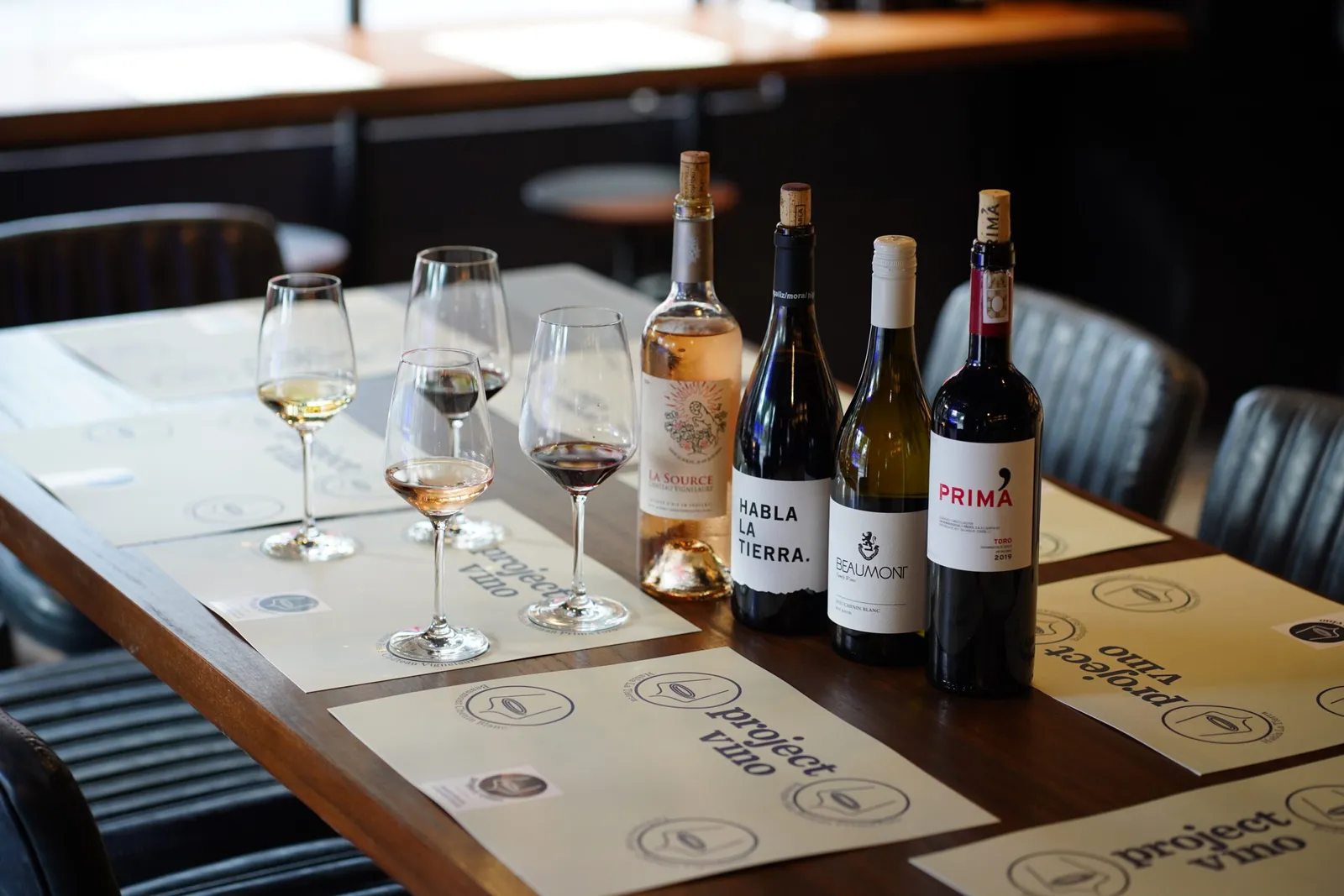 Four wine bottles and four poured glasses set on a Project Vino dining table.