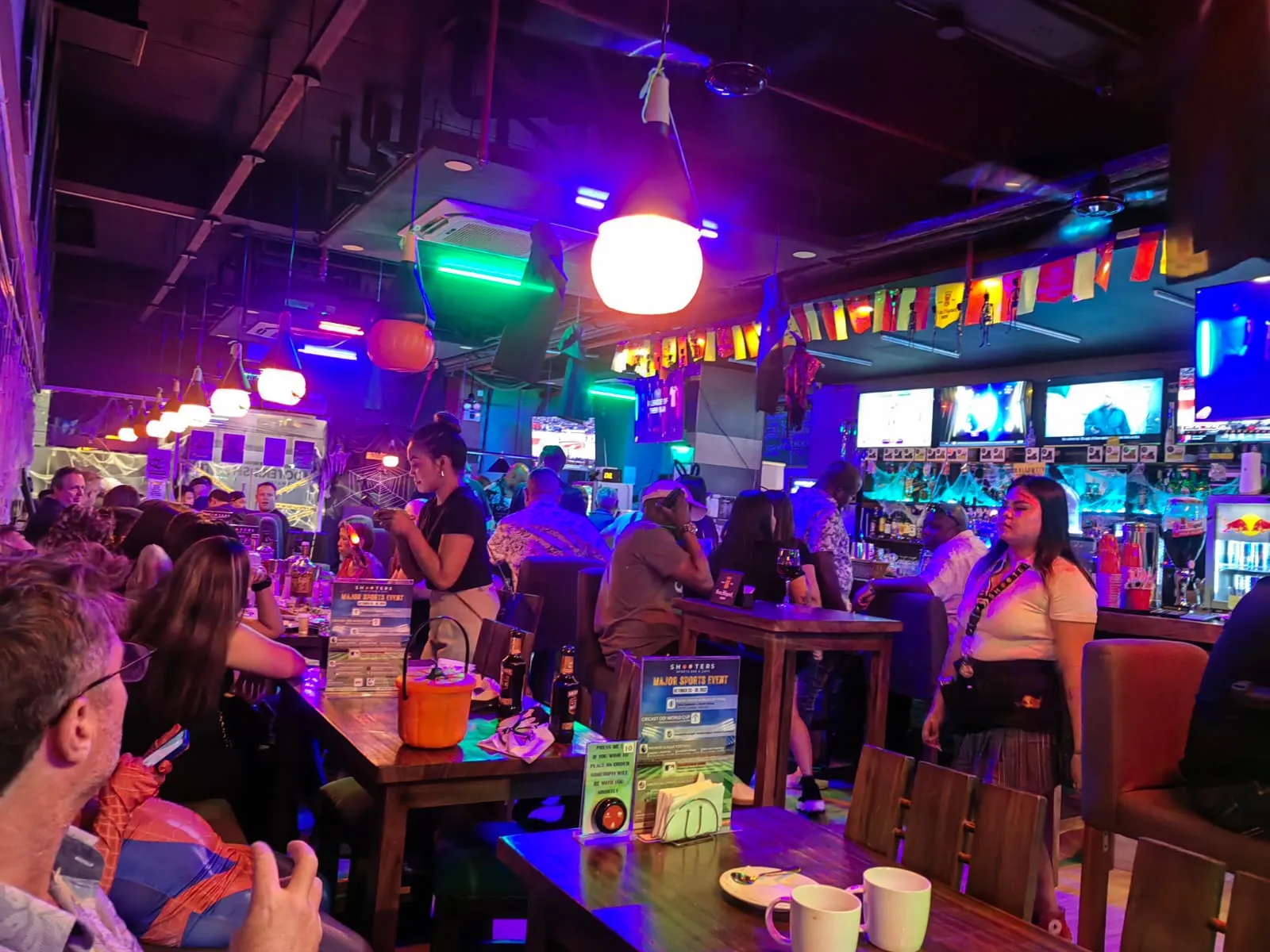 Busy sports bar room under coloured lights with several screens above the counter