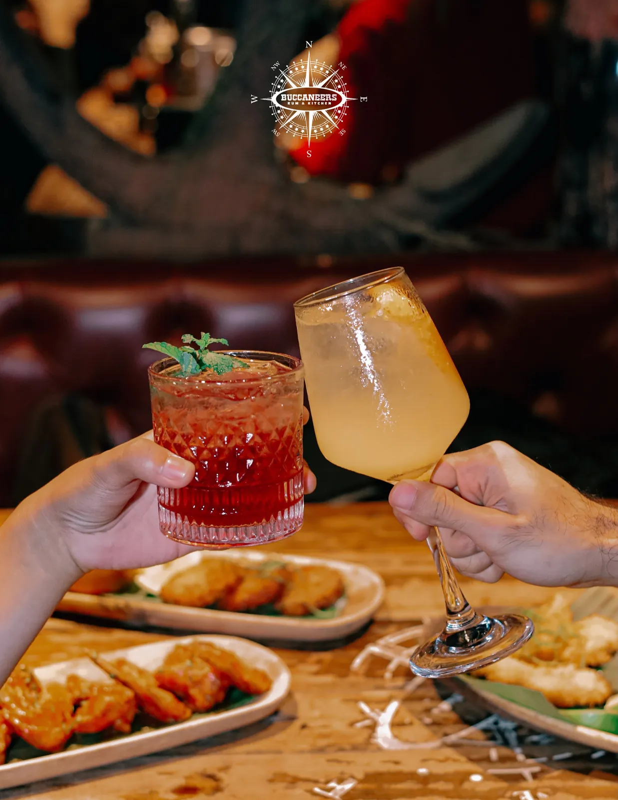Two cocktails held above plates of glazed wings inside the bar.