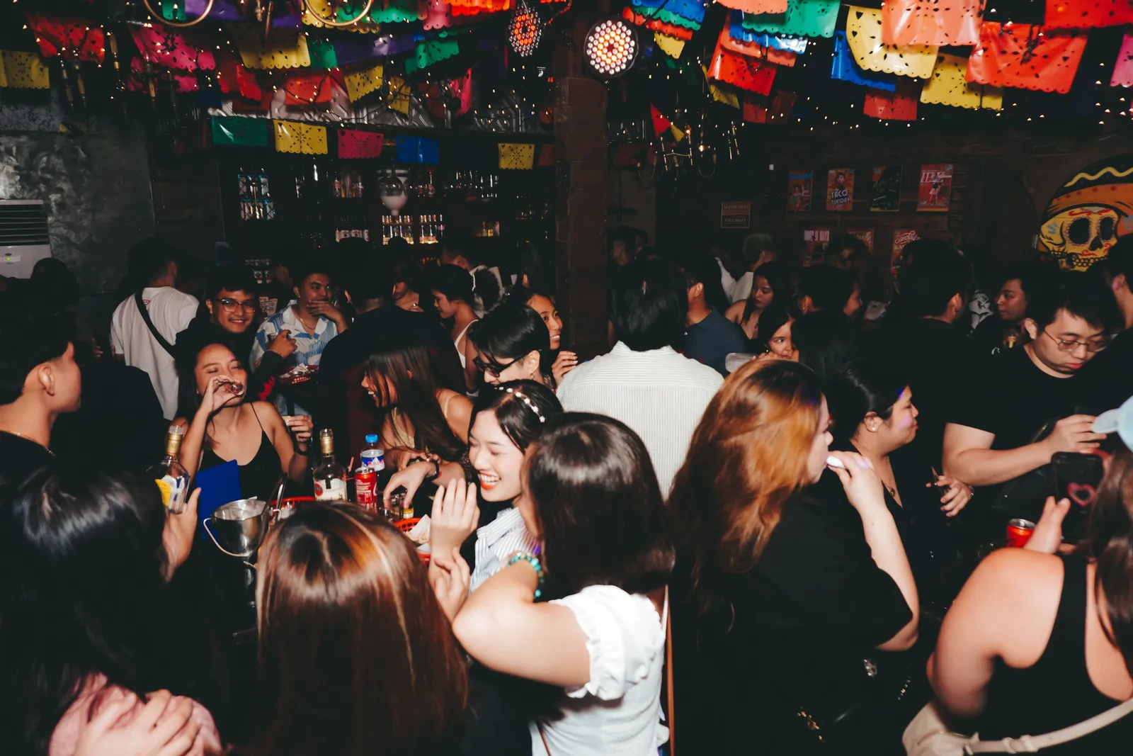 A packed Almacen room at night beneath colorful papel picado and spotlights.