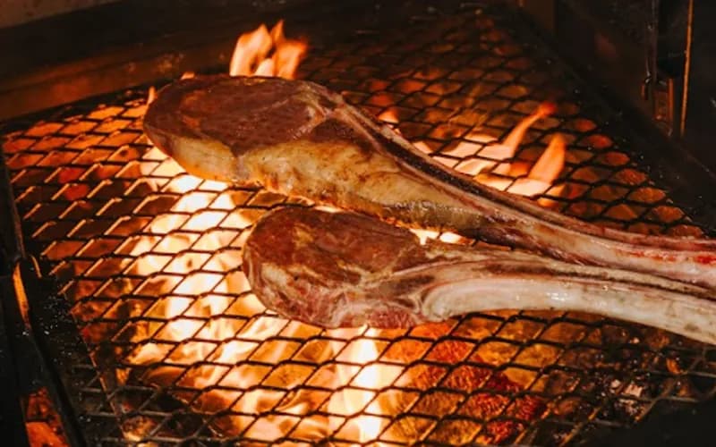 Bone-in steaks cooking over flames on the firewood grill.